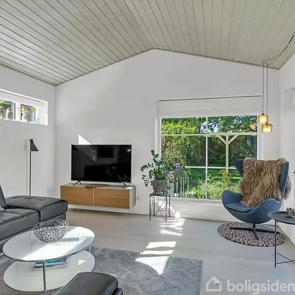 Stue med sort lædersofa og farverig pude, fladskærms-TV på vægskab. Store vinduer giver lysindfald og udsigt til haven. Moderne indretning med planter og designsstol i hjørnet.