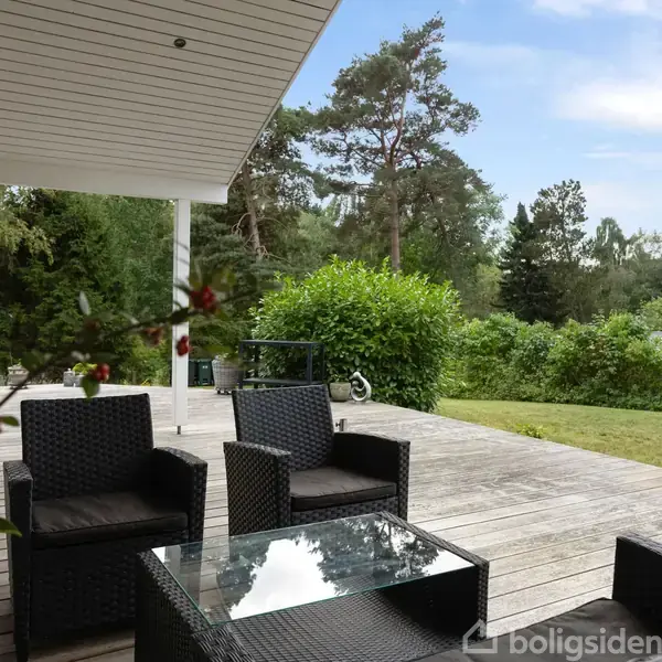 Terrassemøbler i sort flet står stille på en træterrasse med et glasbord, omkranset af en frodig have med græsplæne og buske, under en delvist overdækket himmel.