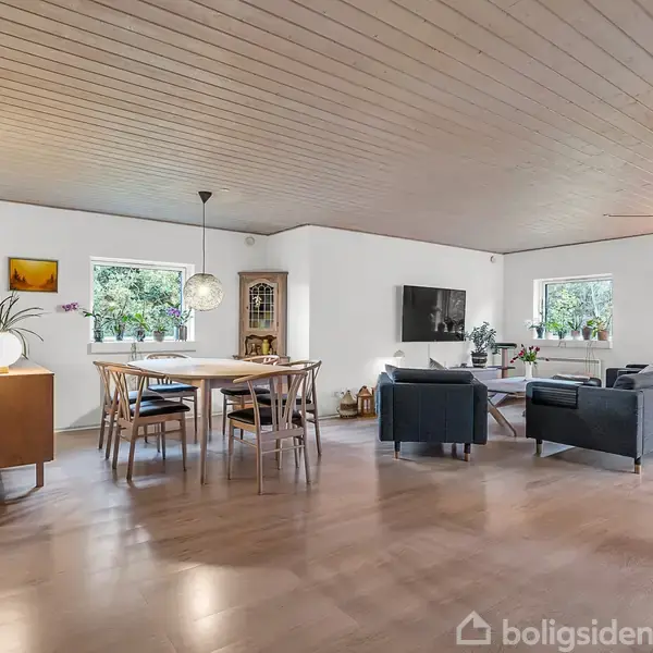 En moderne stue med trægulv indeholder et spisebord med stole midt i rummet. I baggrunden er en sofa og TV. Store vinduer giver naturligt lys i rummet.