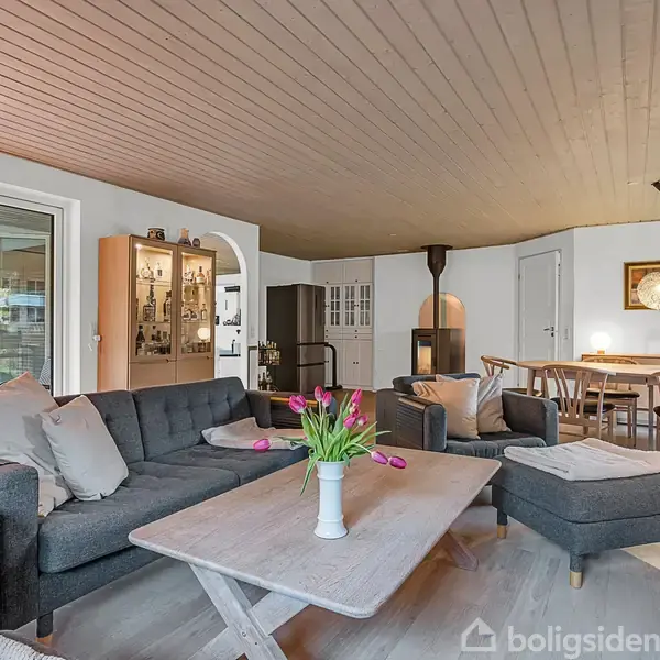 Vase med lyserøde tulipaner står på sofabord foran gråt sofaarrangement. I baggrunden ses spisestue med langbord, hængende lampe og brændeovn, belyst af naturligt lys fra vinduerne.