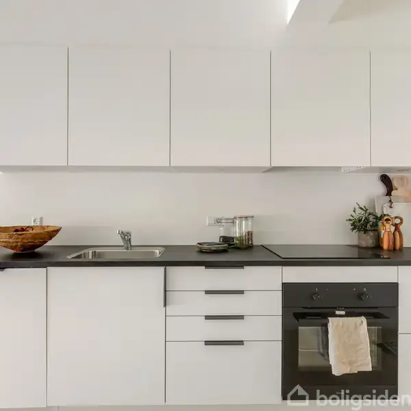 Hvidt køkken med flade skabe og sort bordplade, funktionelt arrangeret med vask, ovn, og dekorativ kurv. En plante og krydderier tilføjer liv til det minimalistiske miljø.