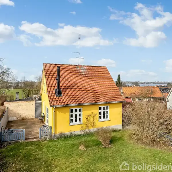 Gult hus med rødt tegltag, beliggende i en have med græs og buske. Træterrasse til venstre og naboer med lignende huse i baggrunden under en blå himmel med skyer.