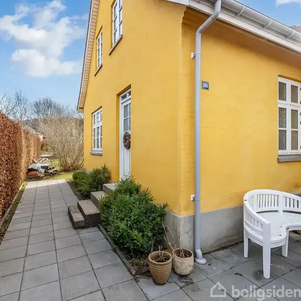 Gulmalet hus nær hæk, hvor der står to små krukker og en hvid havestol på en flisebelagt terrasse. Skyfri himmel skaber en lys og indbydende atmosfære.