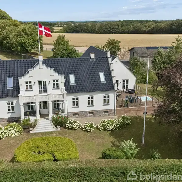 En stor, hvid villa med flagstang bærer Dannebrog. Den ligger i en have med blomsterbede forrest og er omgivet af marker og skov i baggrunden under en blå himmel.
