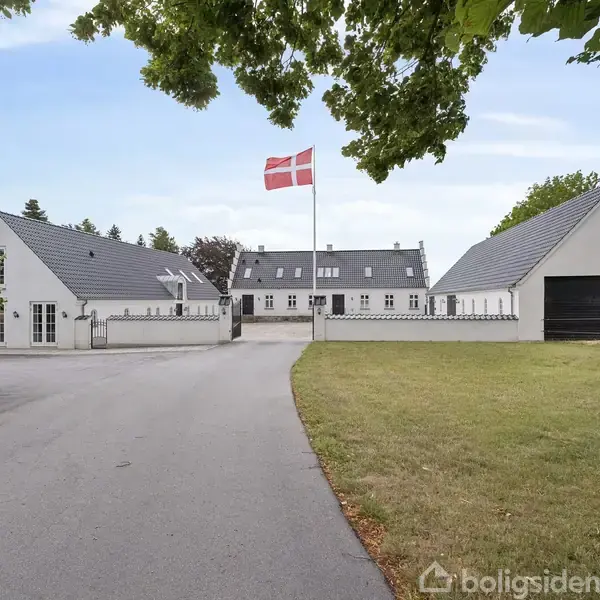 Et landhus med hvid facade og grå tage står ved en indkørsel omgivet af græsplæner. Et dansk flag vajer på en flagstang i midten.