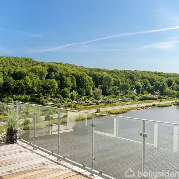 En balkon med glasrækværk og en plante i krukke står overfor en sø. I baggrunden ses en skovklædt bakkeskråning og en klar blå himmel.