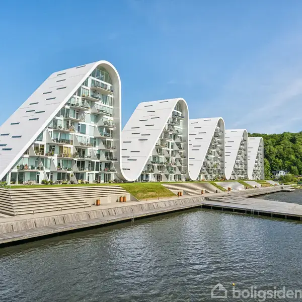 Bølgelignende bygninger står langs en vandkant, med frodige træer i baggrunden og en blå himmel. De karakteristiske hvide strukturer reflekterer i vandet foran dem.