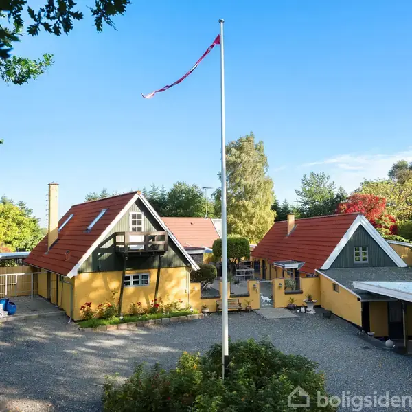 Gul gård med to bygninger, omgivet af træer. En flagstang med Dannebrog står i midten af gårdspladsen. Solrigt vejr og blå himmel.
