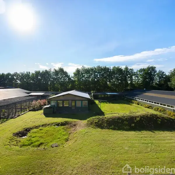 En enkeltstående bygning står i midten af en græsplæne med træer i baggrunden. Bygningen er omgivet af store strukturer på begge sider under en klar blå himmel.