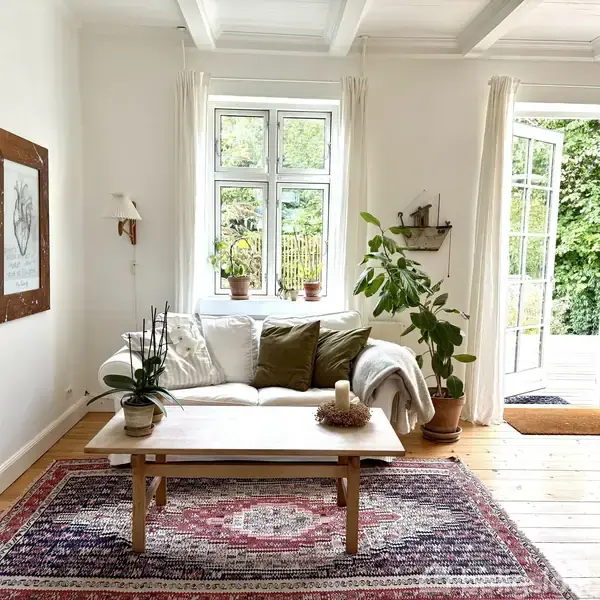 Sofa med puder står foran et stort vindue med gardiner, omgivet af grønne planter. Et træbord med lys står på et farvet tæppe. Dør fører ud til en have.