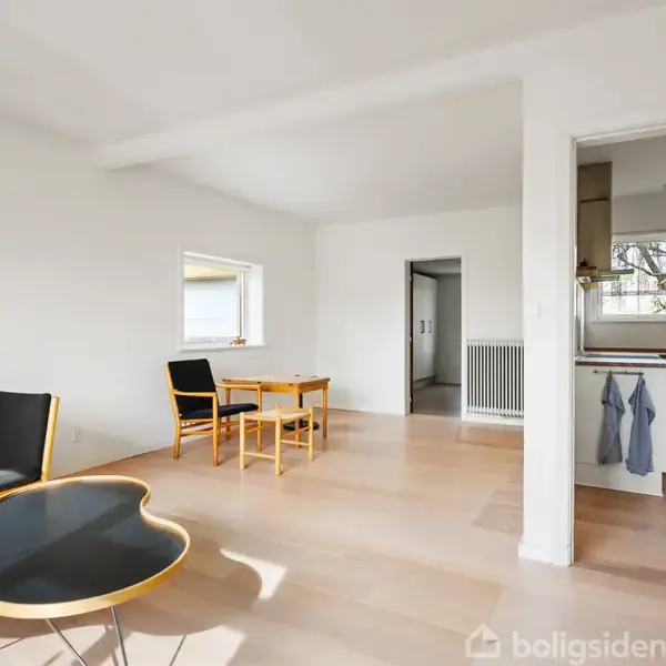 En stue med en sort sofa med en farverig pude, et buet sofabord og et lille spisebord med to stole. I baggrunden ses et køkken og en radiator.
