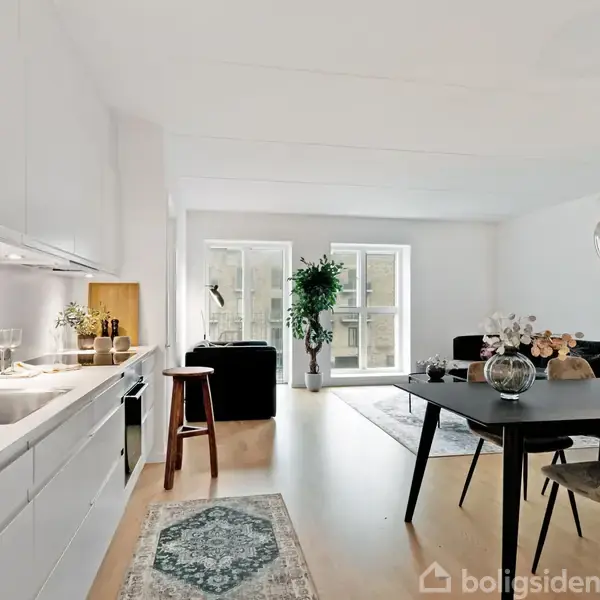 Et moderne køkken-alrum har hvide skabe og vask; spisebordet står ved siden af en hængende lampe. I baggrunden ses en stue med sorte sofaer og store vinduer.