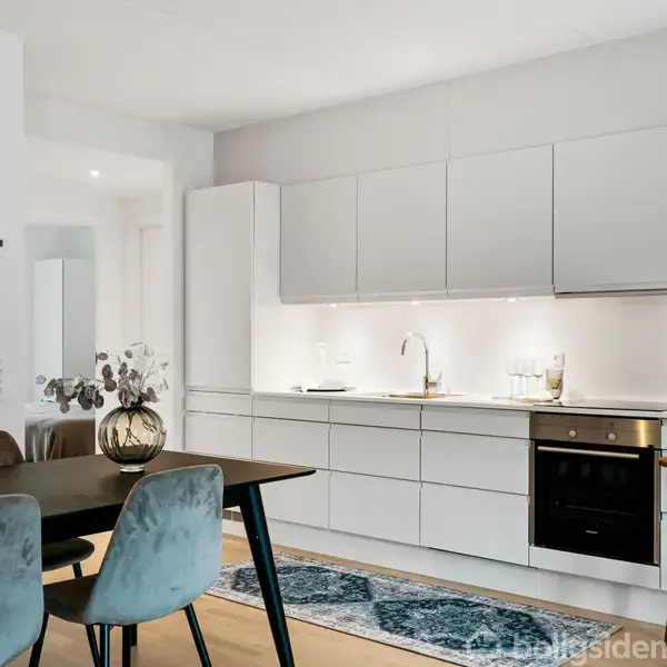 Et moderne køkken med hvide skabe og en sort ovn integreret i væggen. Et træbord med blå stole er placeret i forgrunden. Enkelt belysning og en blomstervase dekorerer rummet.