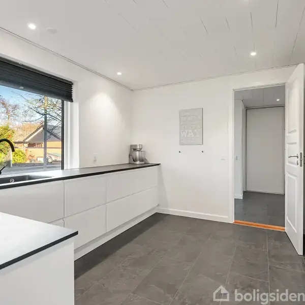 Et moderne køkken med hvide skabe og sort bordplade. En vask med vandhane ved et stort vindue viser en have. Til højre er en åbentstående dør til en anden gang.