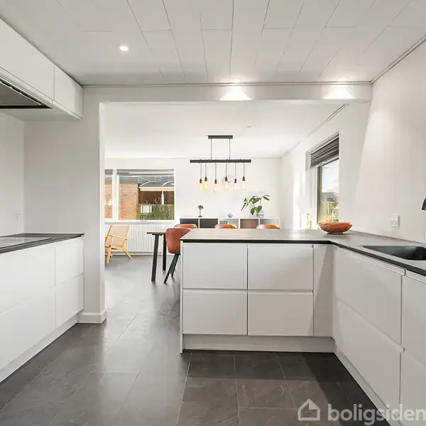 Et moderne køkken med hvide skabslåger og sort bordplade er tomt. I baggrunden ses en spiseplads med et træbord og stole. Store vinduer giver lysindfald fra haven.