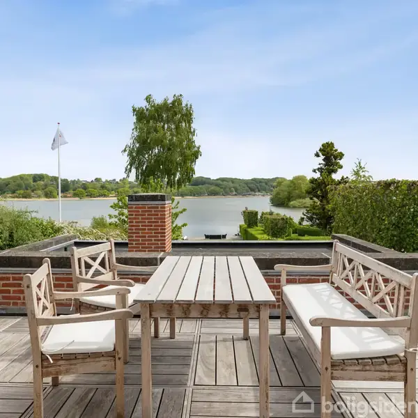 Udendørs terrasse med træmøbler, herunder et bord og stole, står stille. Omkringliggende miljø består af en sø i baggrunden, omgivet af grøn natur og en blå himmel.