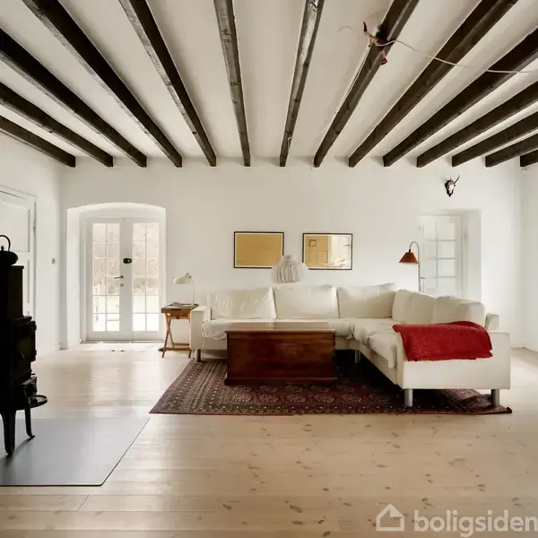 En hvid stue med mørke træbjælker i loftet. En brændeovn står i venstre side. En stor, lys sofa med et rødt tæppe på højre side.