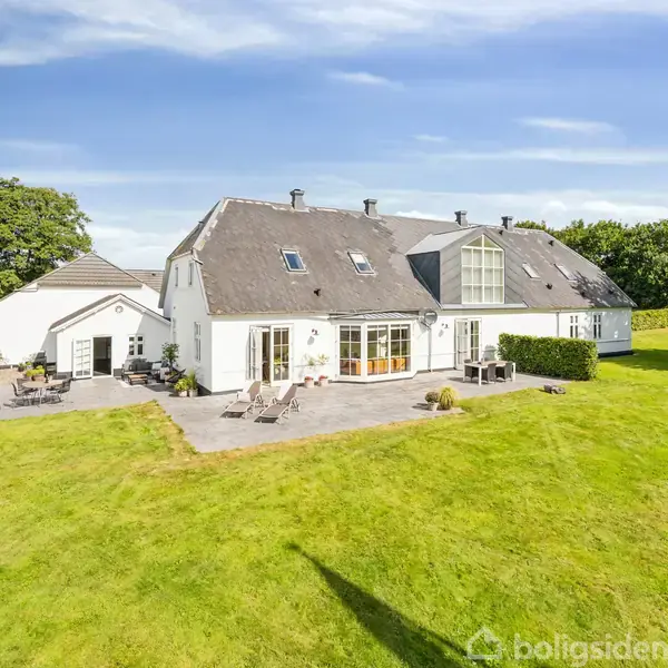 Hus med hvide vægge og grå tag, står stille i en stor grøn have med klare blå himmel og få omgivende træer.