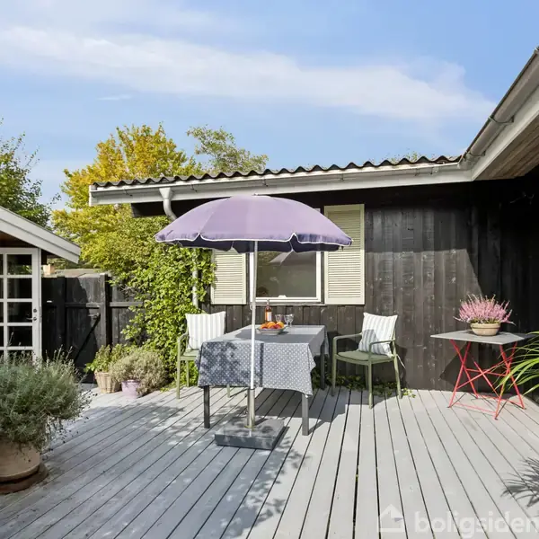 Terrassen har en træbord med en lilla parasol og to stole. En skovgrøn plante står i en krukke. Sortmalet hus omkranser området. Skyfri himmel i baggrunden.