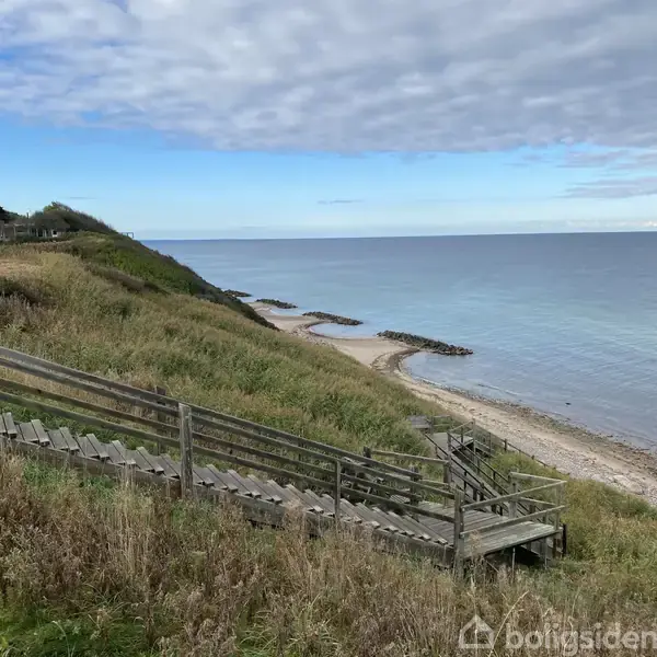En trappe fører ned ad en kystbakke mod en stenstrand. Omgivelserne er præget af græs og sand med horisonten og havet i baggrunden under en skyet himmel.