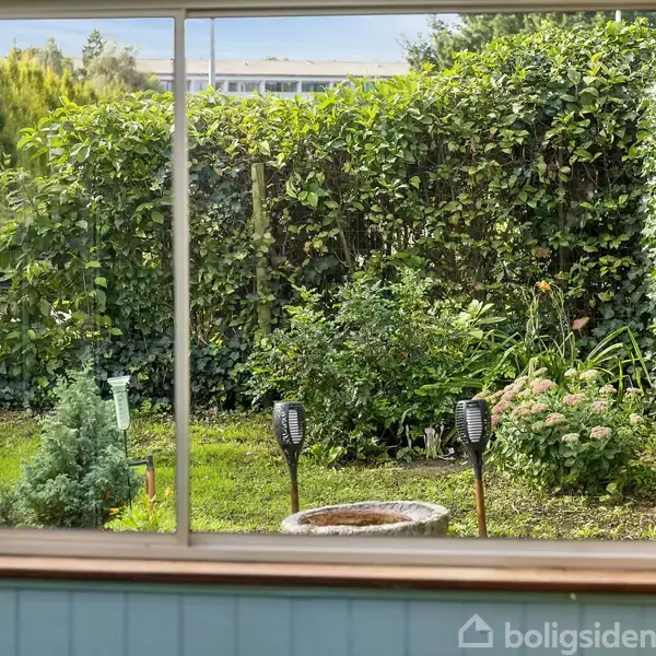 Vinduet viser en have med frodig buskads og græsplæne. Et par solcellelamper står foran buskene, omgivet af små blomster og en lille grøn plante.