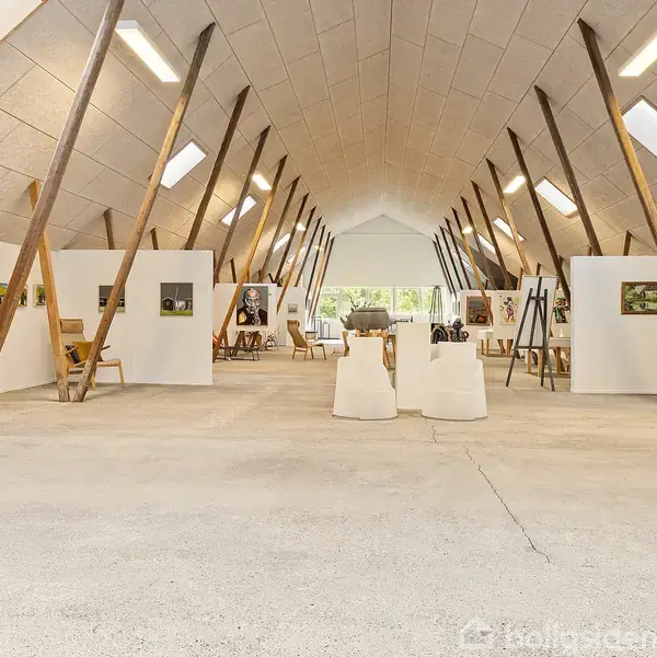Et rummeligt galleri fyldt med kunstværker. Malerier hænger på skillevægge og staffelier, og rummet har et skråt loft med træbjælker og naturligt lysindfald fra ovenlysvinduer.