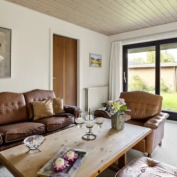 Brun lædersofa og lænestol står omkring et træbord med blomster og stearinlys. Rummet har hvide vægge, kunst på væggene, store vinduer med udsigt til en grøn have.
