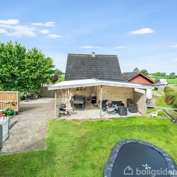 Et hus med skråt tag har en carport foran, hvor der er flere cykler og udendørsmøbler. En trampolin er placeret i en græsplæne, omgivet af grønne marker og træer.