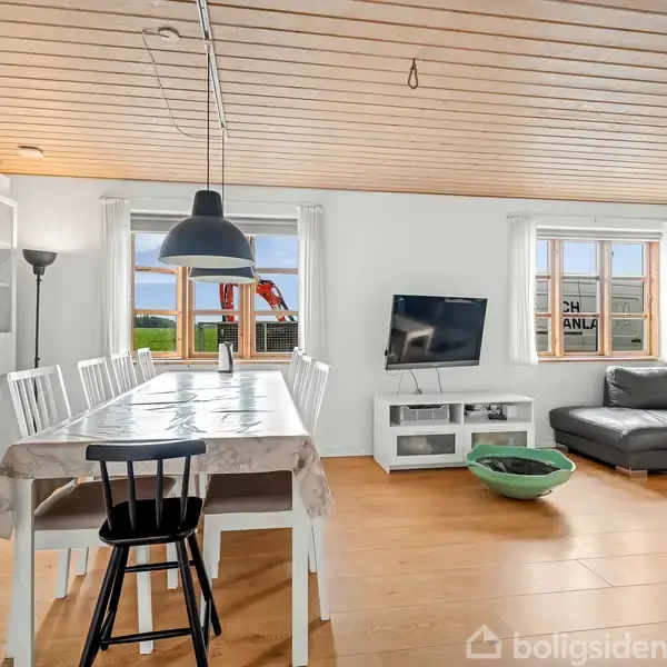 Spisebord dækket med dug og omgivet af stole; ved siden af et hjørnesofaarrangement med sofabord; fladskærms-TV på hvidt møbel under to store vinduer, i et lyst rum.