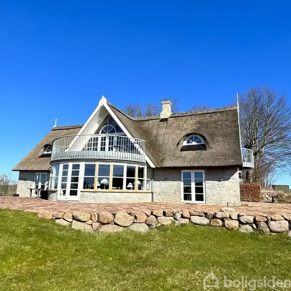 Et stråtækt hus med store vinduer og altan under en klar blå himmel. Foran huset er en grøn græsplæne og en stenkant. I baggrunden ses træer og en lys horisont.