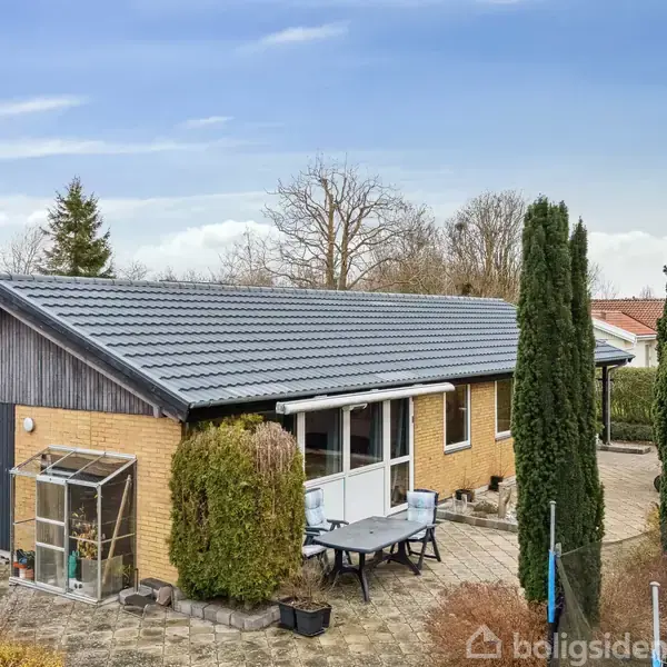 Et gult murstenshus med gråt tag står stille i en have med fliser og træer. Et lille drivhus er ved siden af hoveddøren, omgivet af buske og en terrasse med havemøbler.