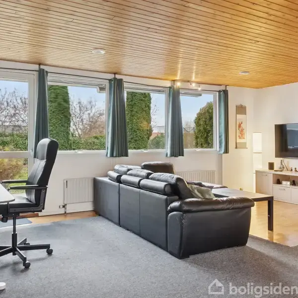 En moderne stue med en sort lædersofa som hovedfokus, orienteret mod et vægmonteret TV og et lavt skab. Rummet har store vinduer med grønne gardiner, der giver udsigt til en have, mens et skrivebord står i hjørnet.