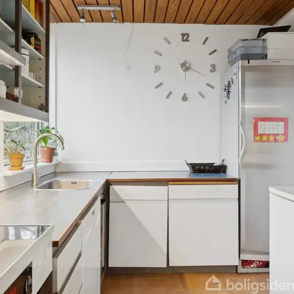 Et moderne køkken med hvide skabe og stålbordplade. Vægur uden urkasse viser tid. Der er en plante i vindueskarm. Køleskab har farverige tegninger på døren.