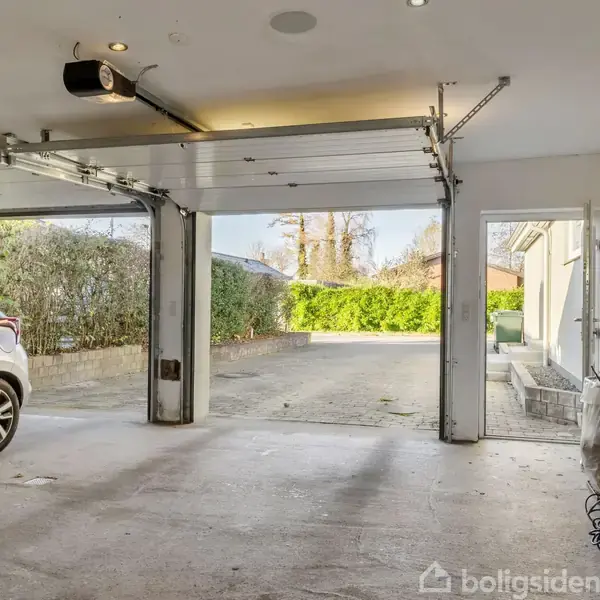 En bil står i en åben garage; dæk er stablet til højre. Udenfor ses en indkørsel med hække, træer og en solrig himmel.