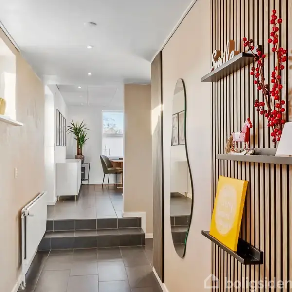 Gang med flisegulv fører til et lyst rum. Væggene er dekorativt panelerede med hylder, spejle, og kunstværker. En plante ses i det fjerne rum.