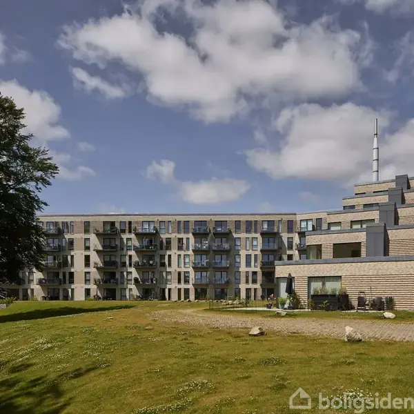 En moderne femetagers boligblok med altaner er omgivet af en græsplæne og et stort træ. Himlen er blå med skyer, og der er ingen synlig tekst.