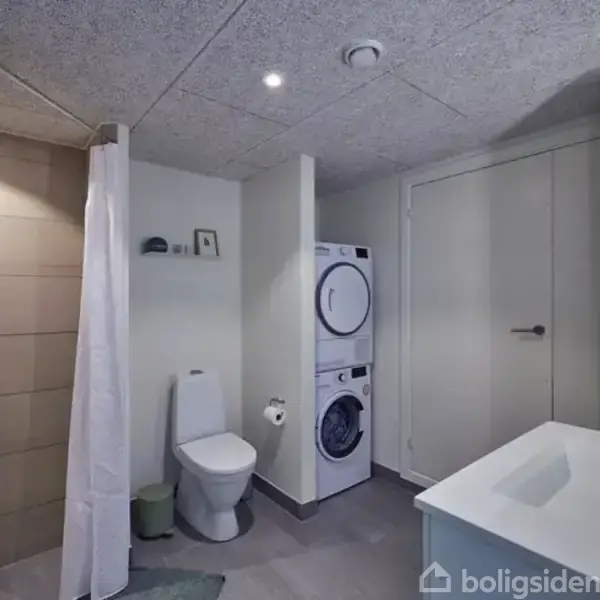 Vaskesøjle står i et moderne badeværelse. Toilettet er ved siden af med en hylde over. Vask med spejl til højre. Flisebelægning på gulv og bruser med forhæng.