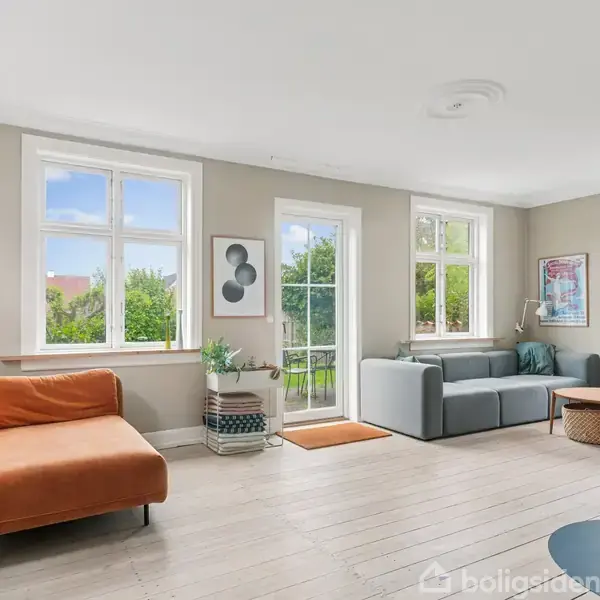 Stue med to sofaer; en orange og en grå, placeret ved store vinduer med udsigt til grønne træer. En moderne pejs står i det ene hjørne. Væggene har moderne kunst.