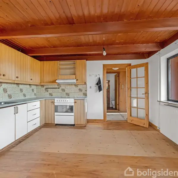 Et køkken med træloft og gulv, hvide skabe og skuffer langs væggen. En ovn står midt i. Et glasdør fører til en lys gang.