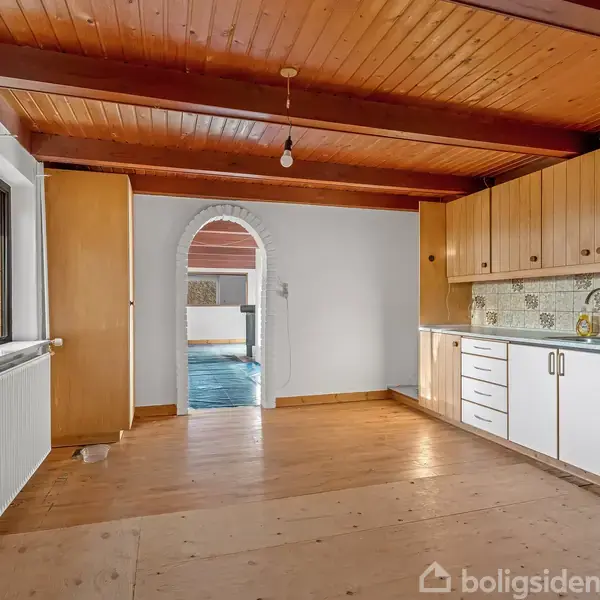 Køkken med hvide skabe langs væggen, ingen apparater synlige, træloft og trægulv. Bueformet åbning fører til andet rum med blå flisegulv. Vindue til venstre med udsigt til træer.