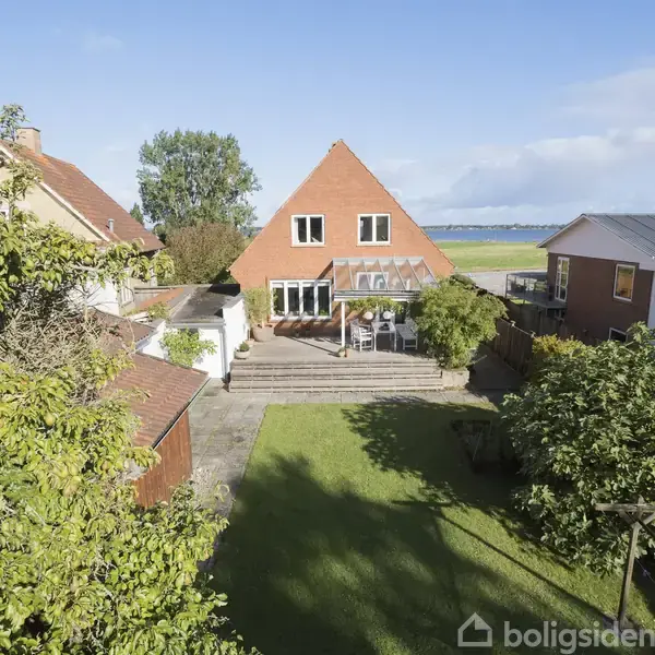 Et rødt murstenshus med en trekantet gavl står stille i en have. Det er omgivet af græsplæne og træer, og der er en terrasse med havemøbler.