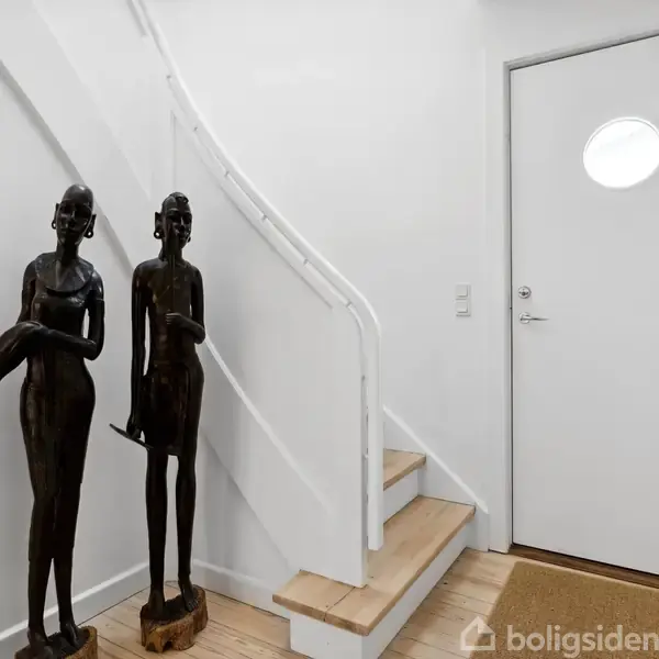 To skulpturer står ved siden af en trappe og en hvid dør med et lille rundt vindue. Miljøet er et enkelt, hvidt rum med trægulv og tæppe.