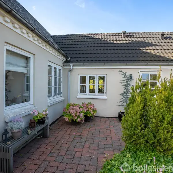 Husfacade med hvide vægge omgiver en flisebelagt terrasse. Potteplanter og buske er placeret omkring området, hvilket skaber en hyggelig atmosfære.