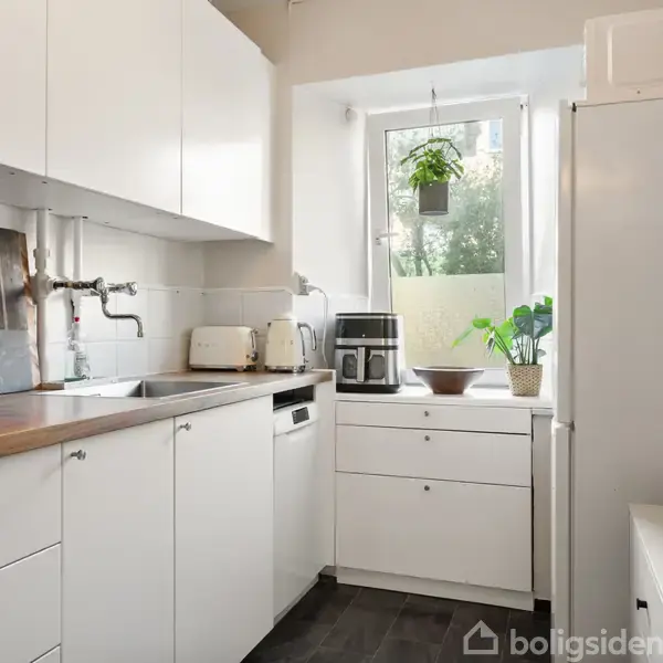 Et lyst køkken med hvide skabe og træbordplade. En vask ved vinduet med planter udenfor. Hvidevarer inkluderer en brødrister og en kaffemaskine. Hylder til højre.