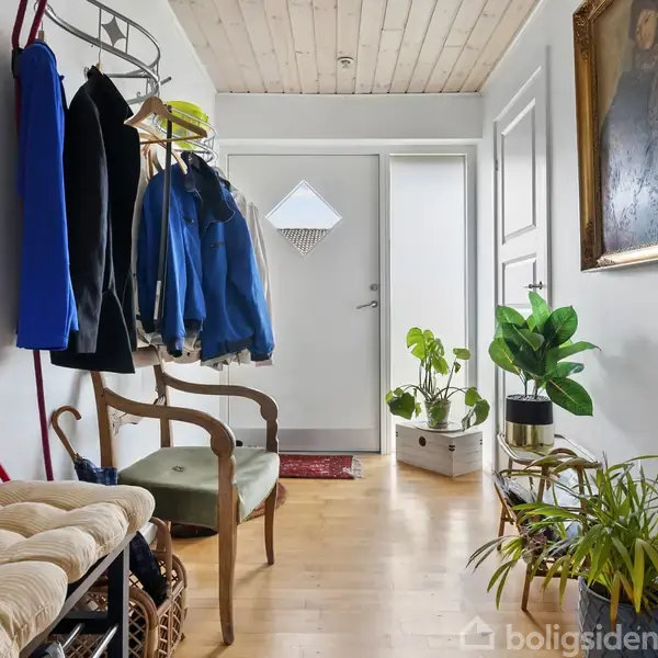 En entré med jakker hængende på et stativ, en stol ved siden af. Trægulv, hvide vægge, planter på gulvet og en maleri hænger på væggen.