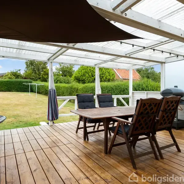 Terrasse med træbord og stole under et gennemsigtigt tag. I baggrunden ses en stor græsplæne omkranset af buske og træer. En grill står ved siden af bordet.