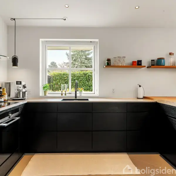 Sort køkken med moderne design, hvor hylder bærer krydderier og pyntegenstande. Gennem vinduet ses grønne buske og et hus. Loftet har minimalistiske lamper. Tekst ignoreret: "Boligsiden".