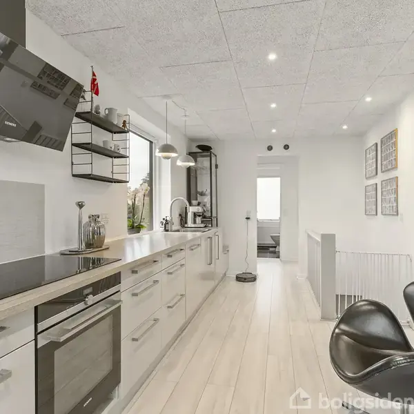 Et moderne køkken med åbne hylder og hvide skabe strækker sig langs væggen. En sort, buet stol står ved siden af. Loftet har indbyggede spots. I baggrunden ses en døråbning.