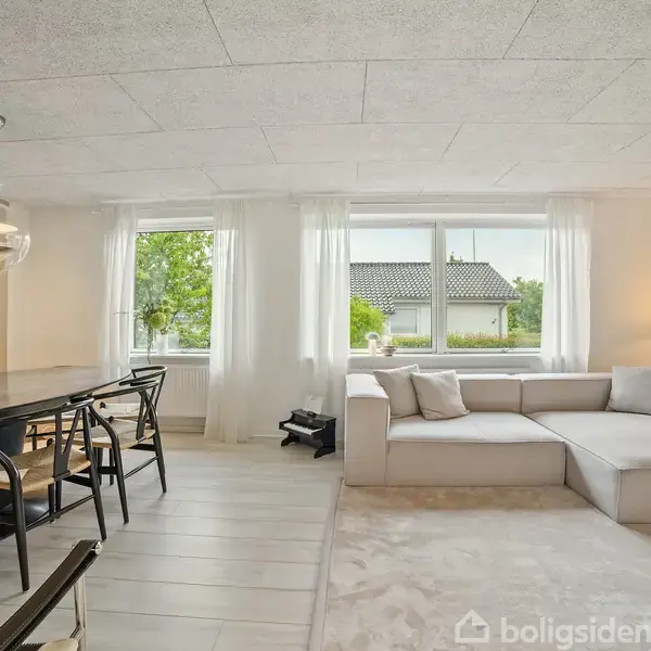 En lys stue med en grå hjørnesofa og en lang spisebord med stole til venstre. Store vinduer med hvide gardiner lukker lys ind, og en lampe tilføjer varme.