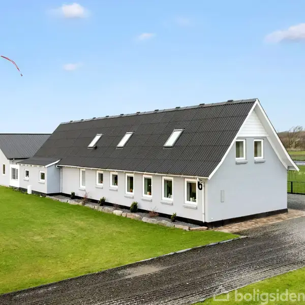 Hvidt, moderne hus med sort tag står på en velplejet plæne. En grusvej fører til huset. En flagstang med flag ses til venstre. Landområde i baggrunden.